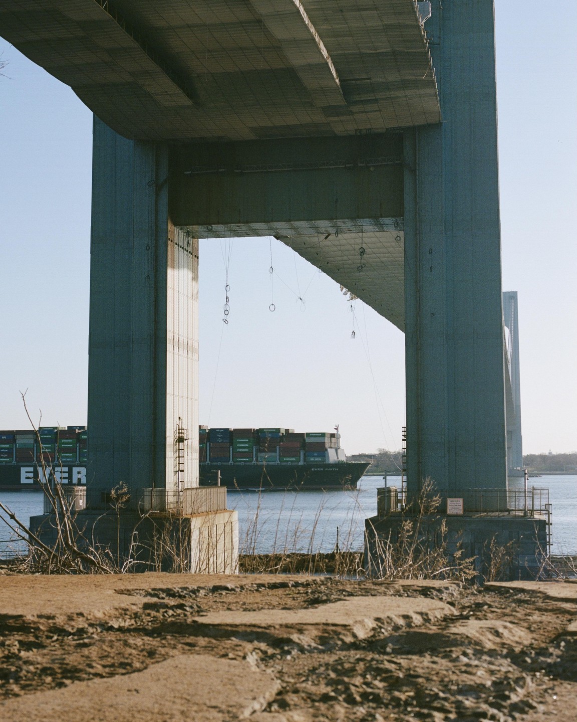 A large cargo ship passes under a massive concrete bridge with a cracked foreground.