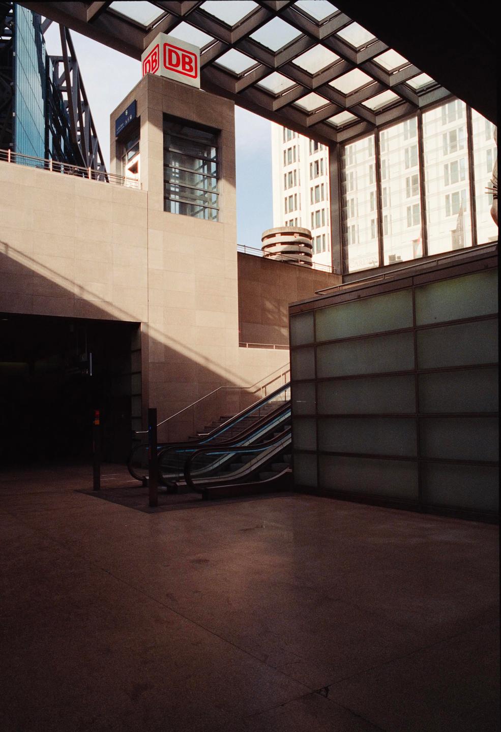 A modern building with a glass roof and escalators is shown with strong shadows.