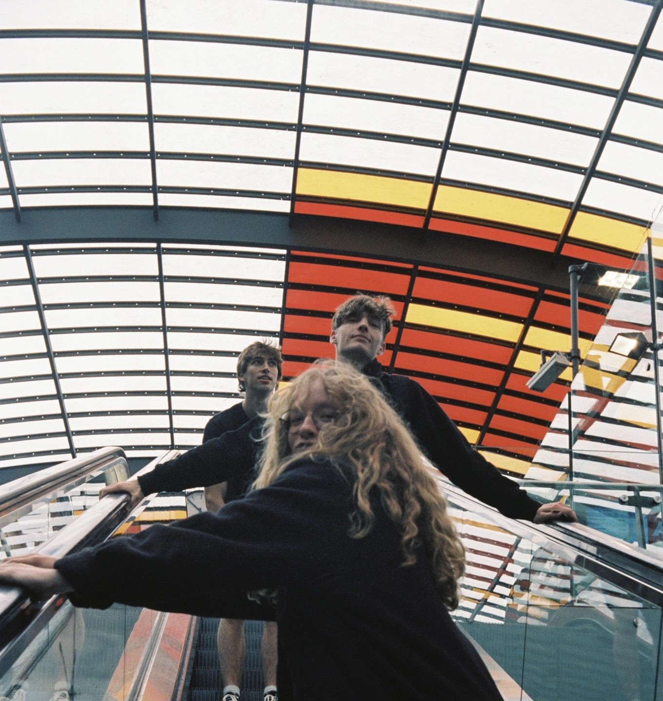 Three young people ride an escalator under a colorful, geometric ceiling.