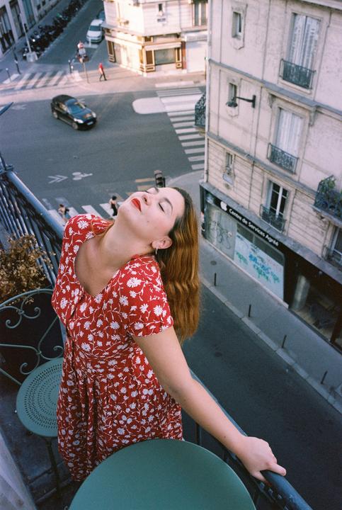 A woman in a red dress with white flowers stands on a balcony overlooking a city street.