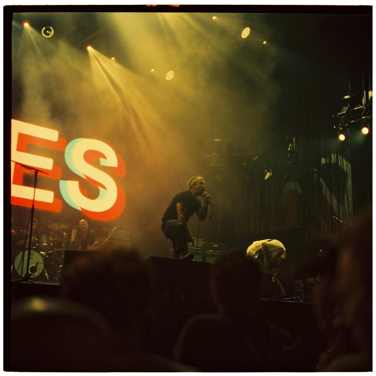 A man sings into a microphone on a stage with large letters spelling "ES" behind him.