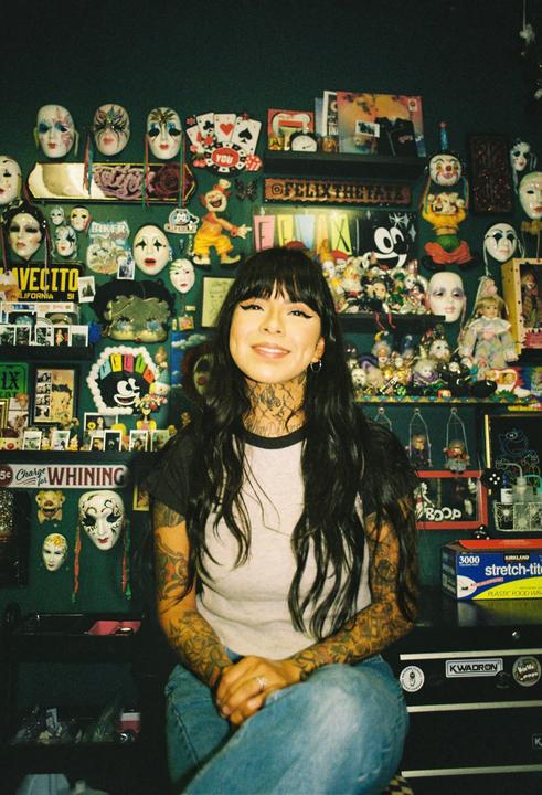 A woman with long dark hair and tattoos sits in front of a wall covered with masks and various objects.