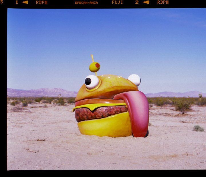 A large burger sculpture with googly eyes and a long tongue sits in a desert landscape under a clear sky.
