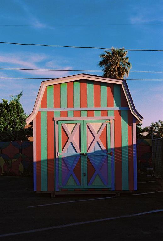 A colorful striped shed with a palm tree and power lines above it stands under a blue sky.