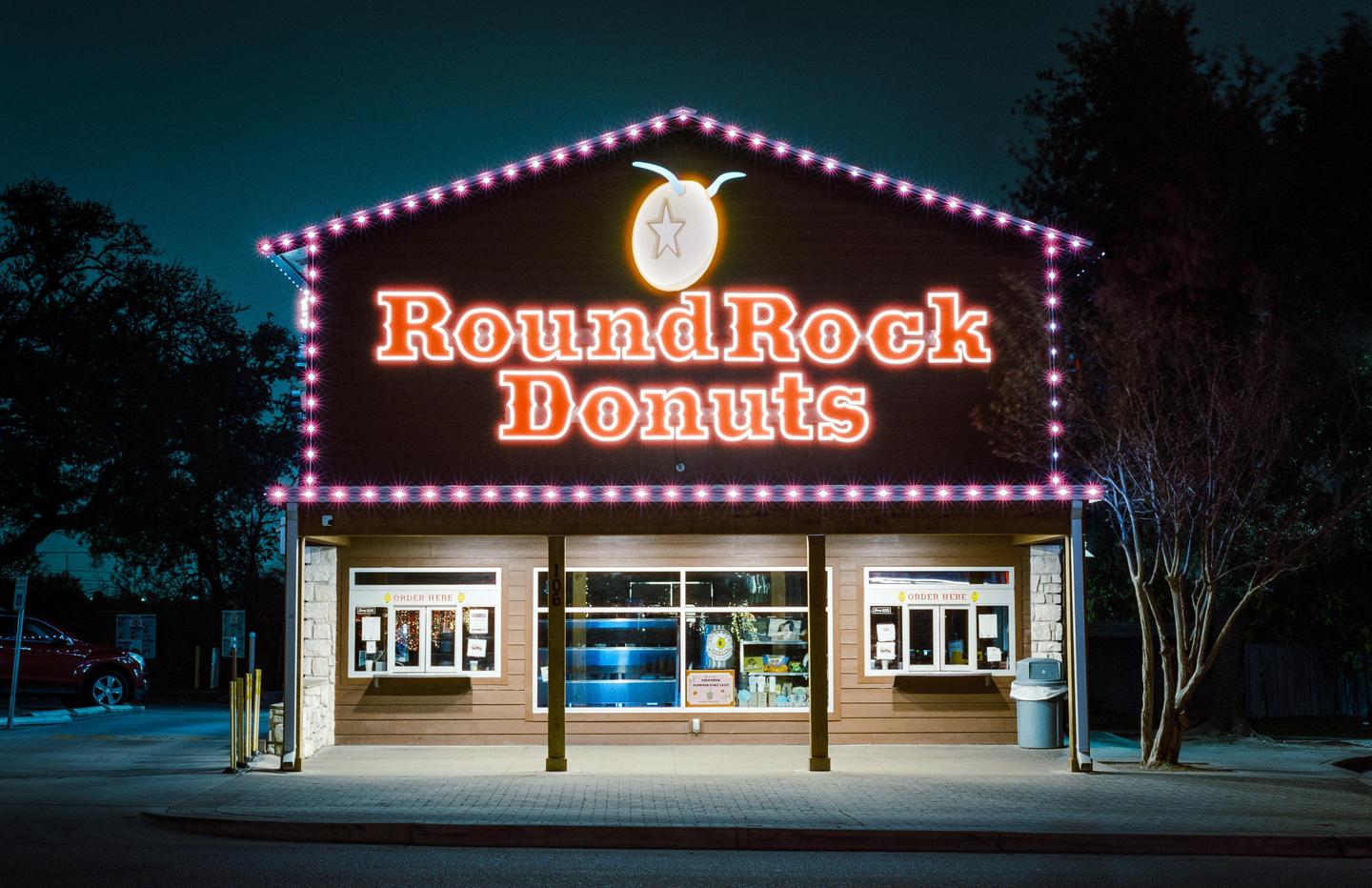 A donut shop with a large neon sign that reads "Round Rock Donuts" is illuminated at night.