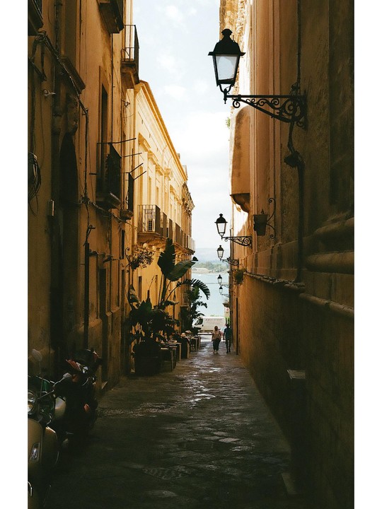 A narrow street with tall buildings on either side leads to a body of water in the distance.