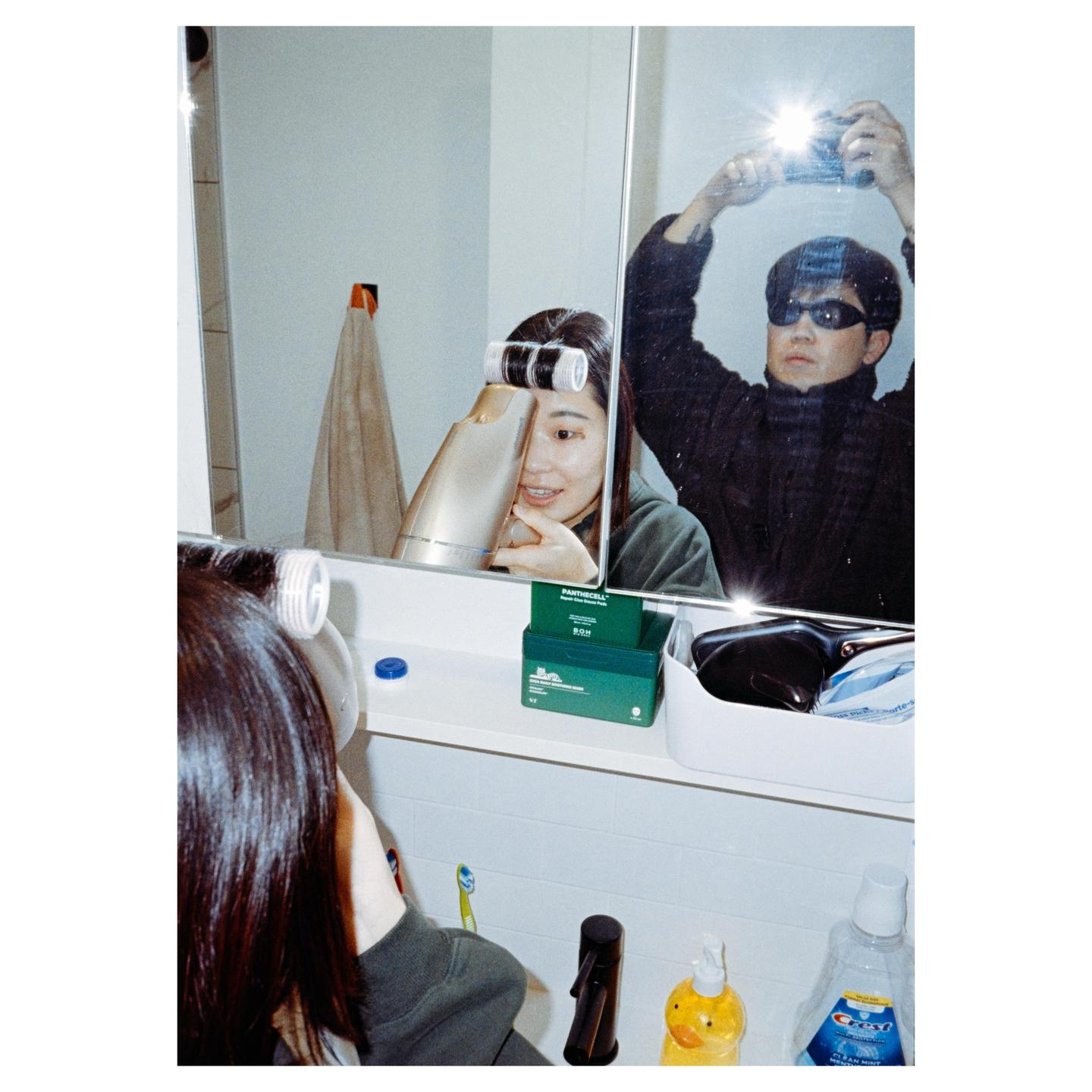 A man takes a mirror selfie of himself and a woman who is styling her hair in a bathroom.