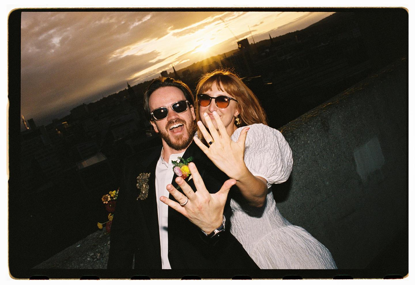 A couple wearing sunglasses shows off their rings at sunset with a city in the background.