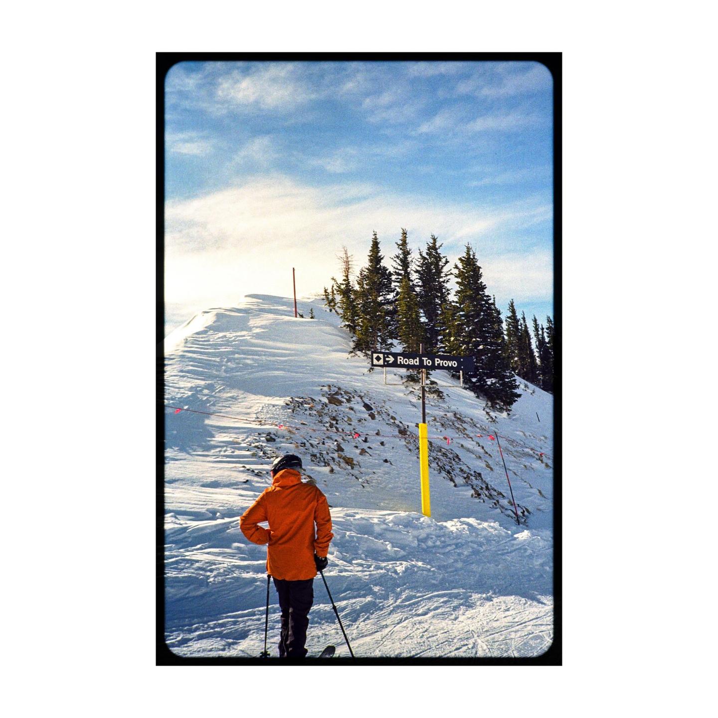 A skier in an orange jacket stands on a snowy mountain slope with pine trees and a sign pointing to Provo.