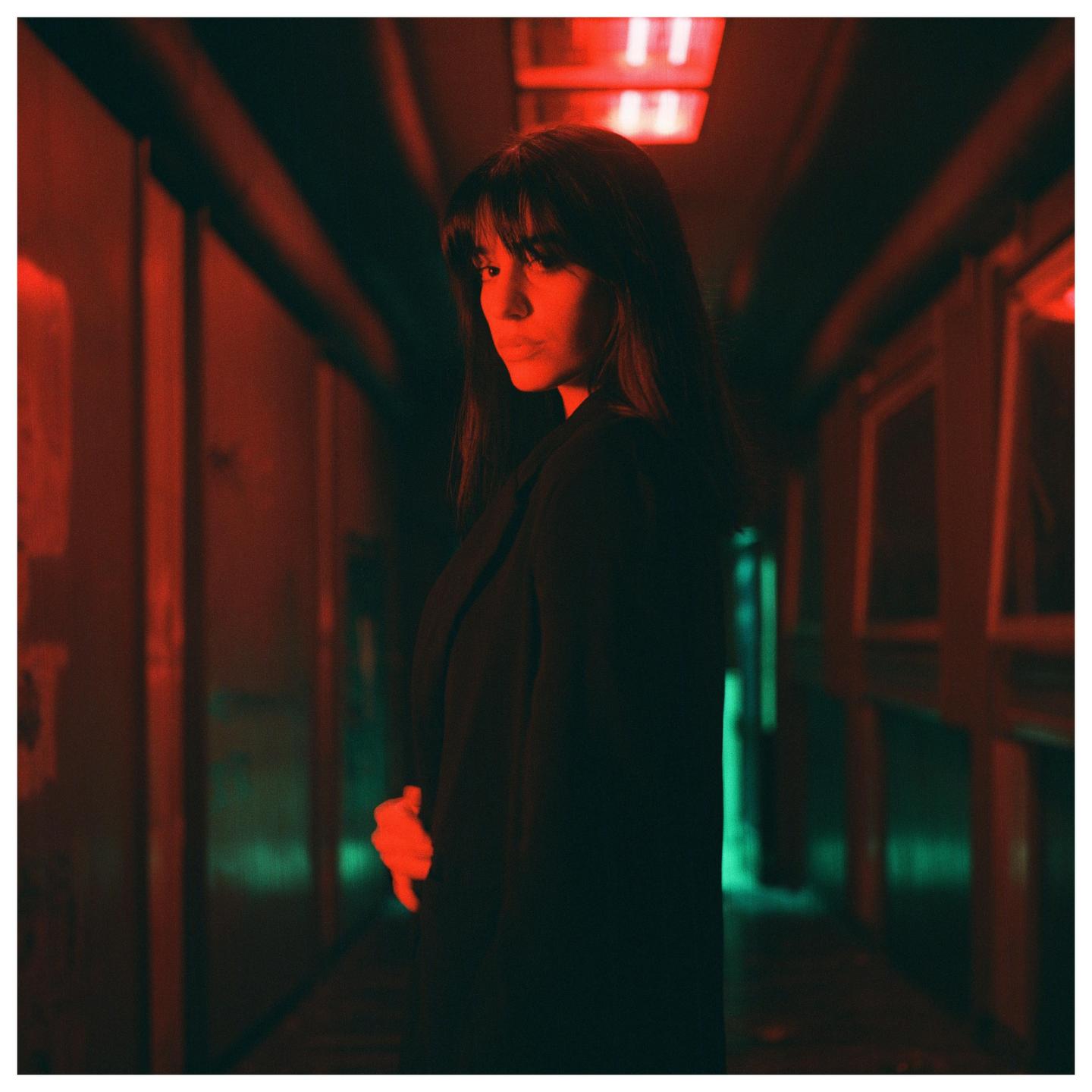 A woman with long hair and bangs looks over her shoulder in a red and green lit hallway.