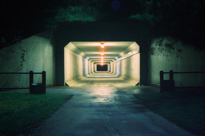 A long, illuminated tunnel stretches into the distance at night, with grass and a barrier on either side of the entrance.