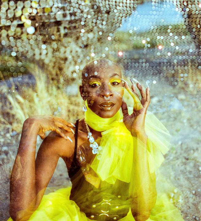 A woman with yellow eye makeup and a yellow scarf sits in a field with a sparkly overlay.