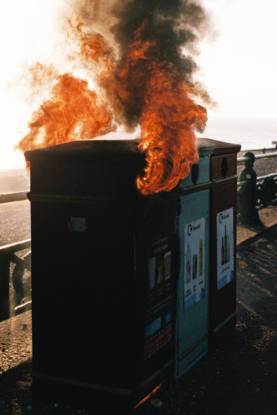 Flames and smoke rise from a burning trash can next to a recycling bin on a sunny day.