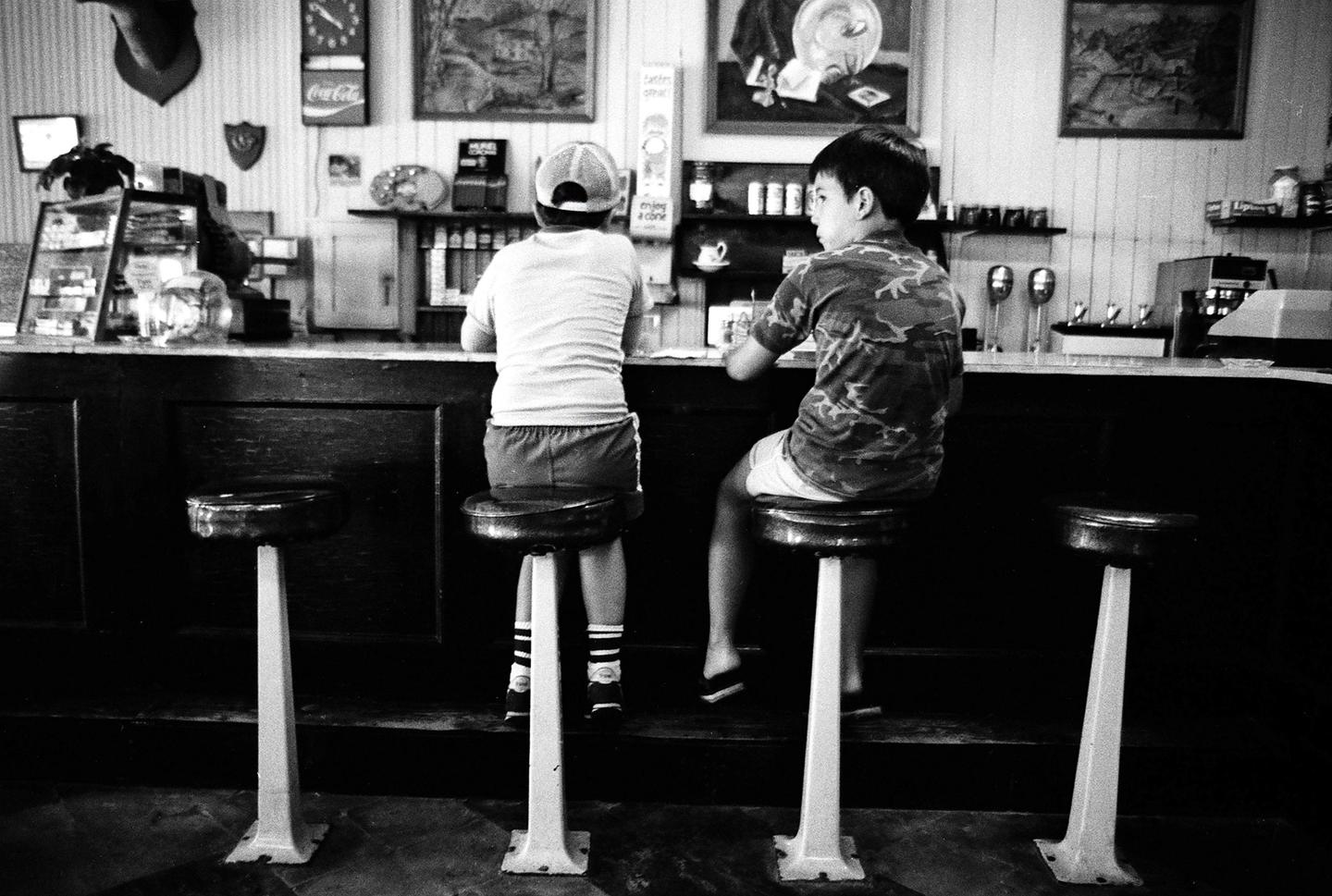 Two boys sit at a counter in a diner with various items on the wall and counter behind them.