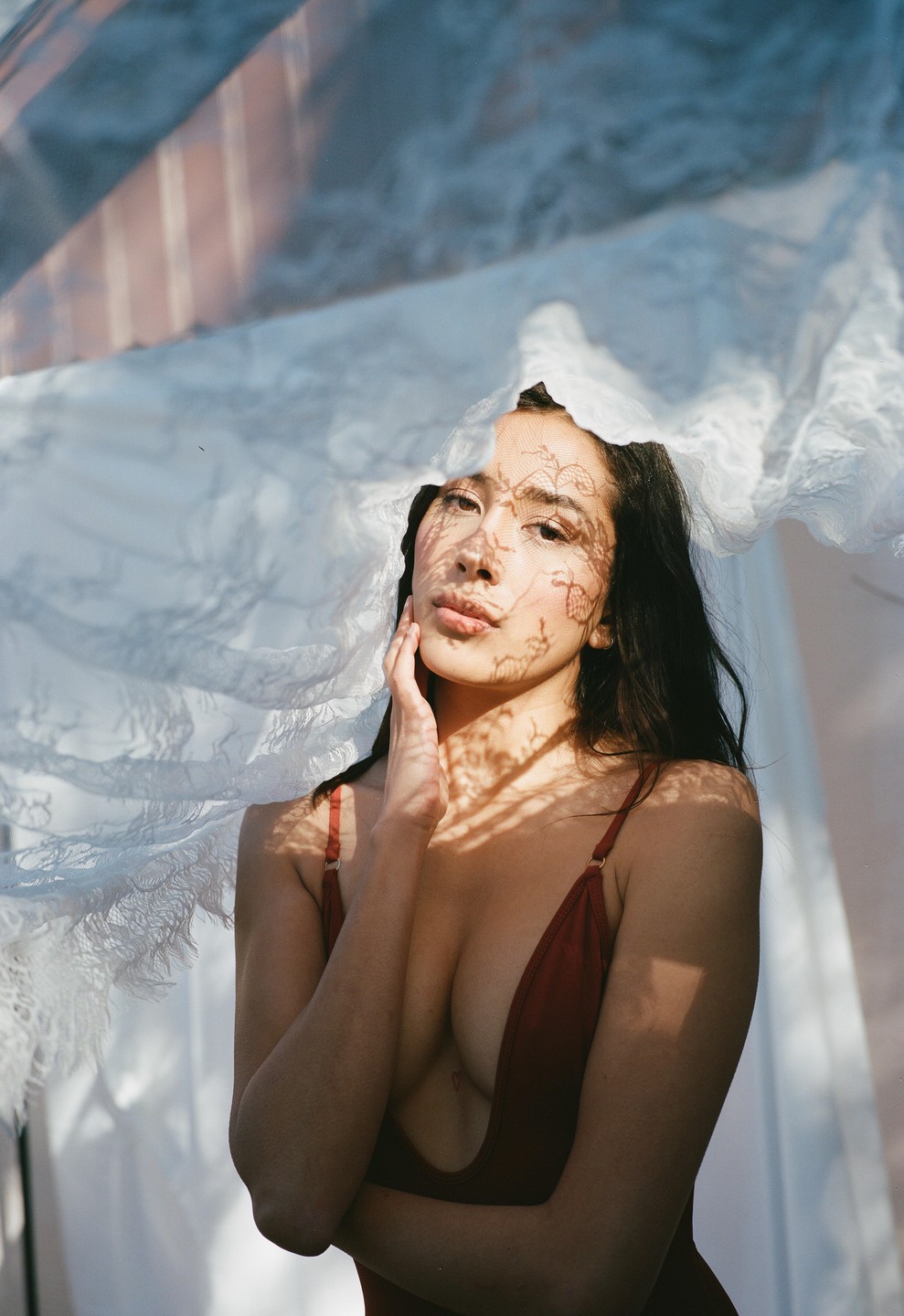 A woman in a red top is partially covered by a white lace fabric, with shadows cast on her face and body.