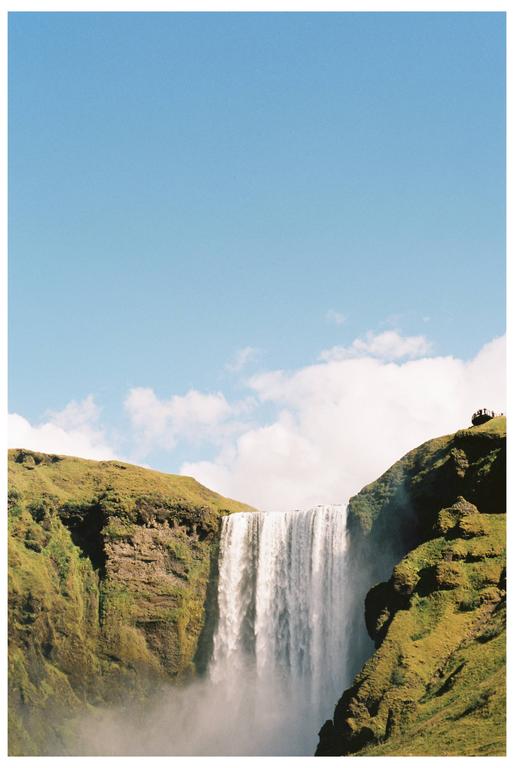 A powerful waterfall cascades between two green, rocky cliffs under a bright blue sky with scattered clouds.