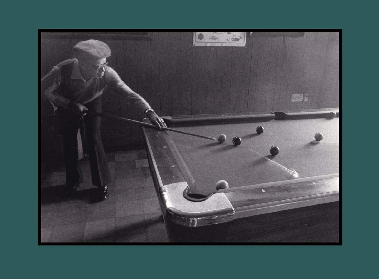 A man in a flat cap leans over a pool table, cue stick in hand, aiming at a cue ball.