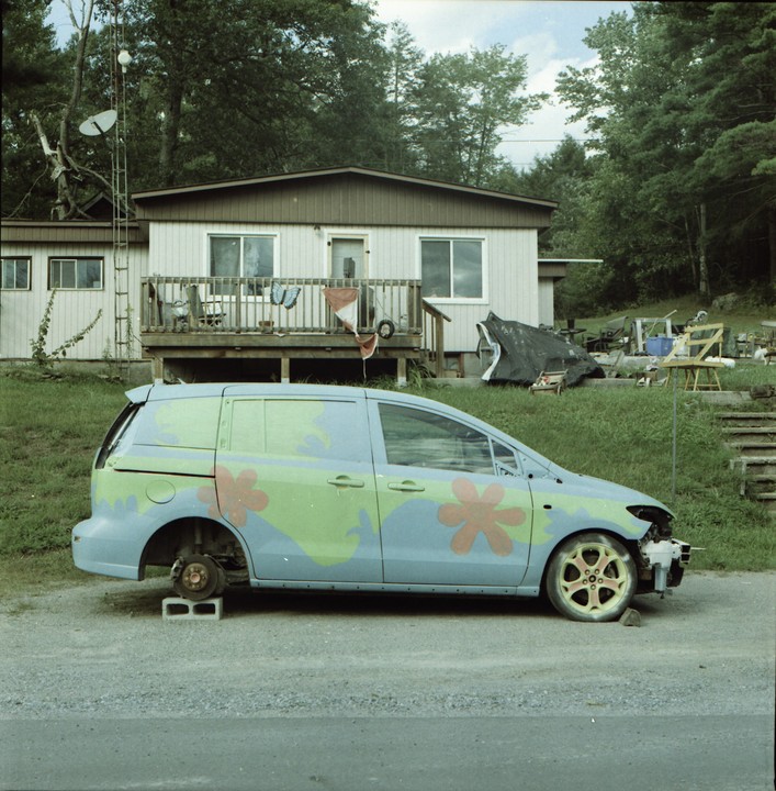 A light blue van with green and orange flower patterns is parked on a gravel road in front of a house with a wooden porch.