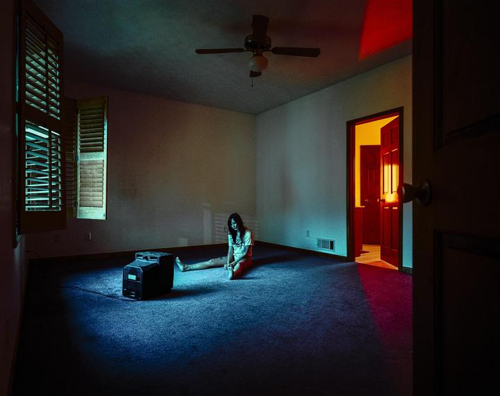 A woman sits on the floor next to a television in a room with a ceiling fan and open doors.
