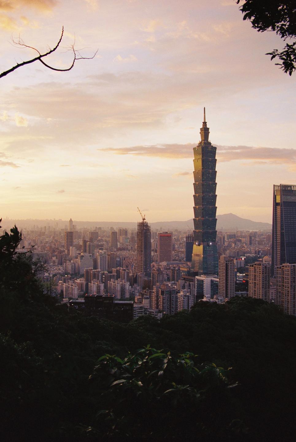 A tall skyscraper rises above a city skyline at sunset, viewed from a hillside with dark foliage in the foreground.
