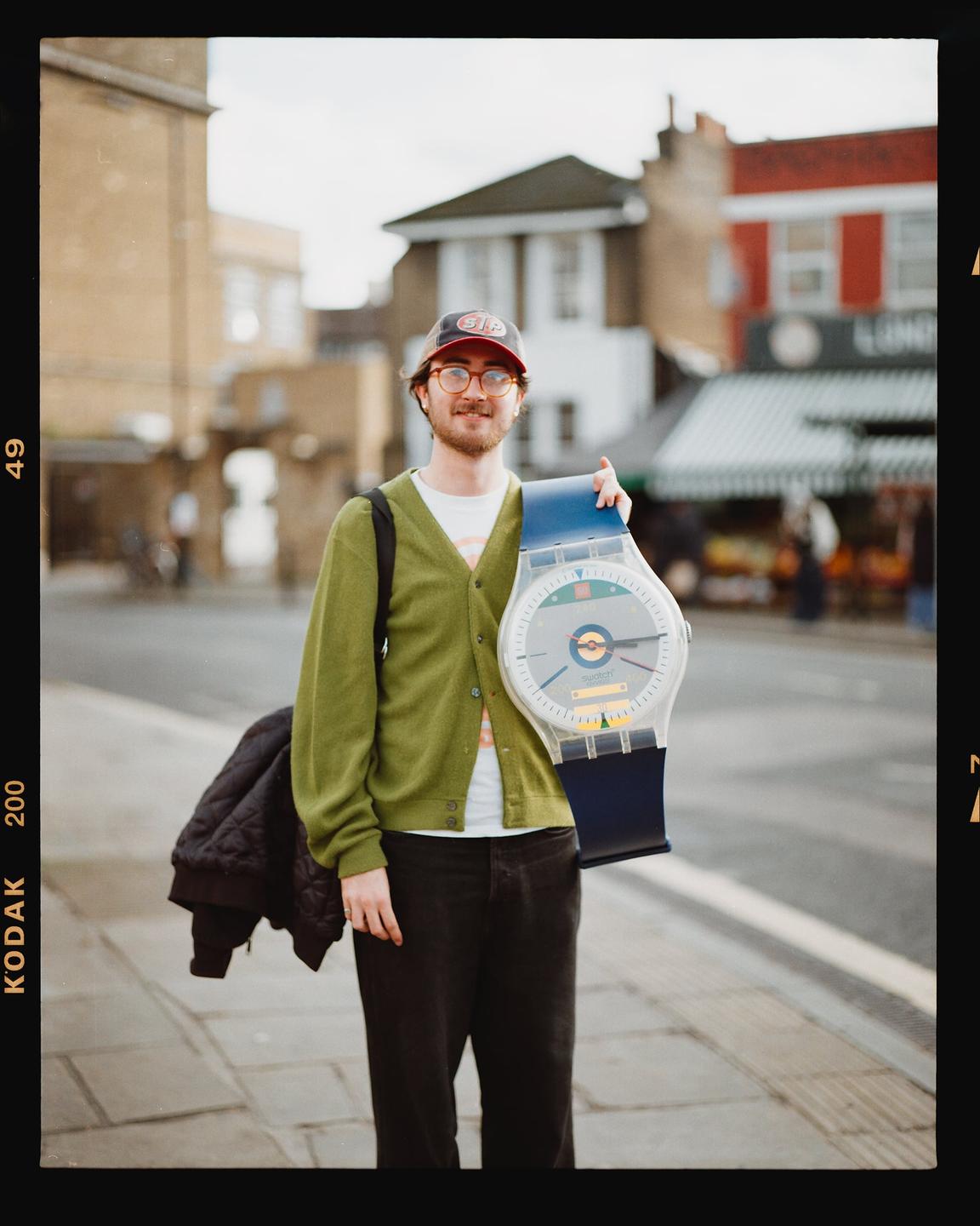 A man wearing a green cardigan and a baseball cap stands on a sidewalk holding a large novelty watch.
