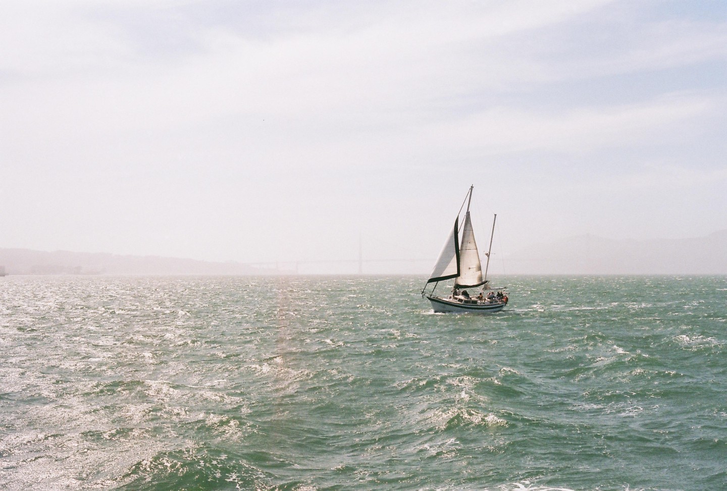 A sailboat with people on board sails on choppy water with a bridge in the distance.