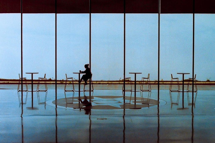 A woman sits at a high-top table with a drink, silhouetted against a large window with a blue sky outside.
