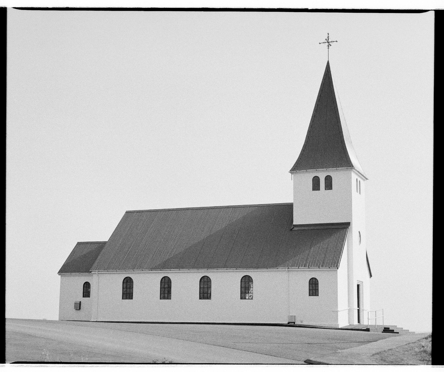 A white church with a tall steeple stands on a slight incline under a bright sky.