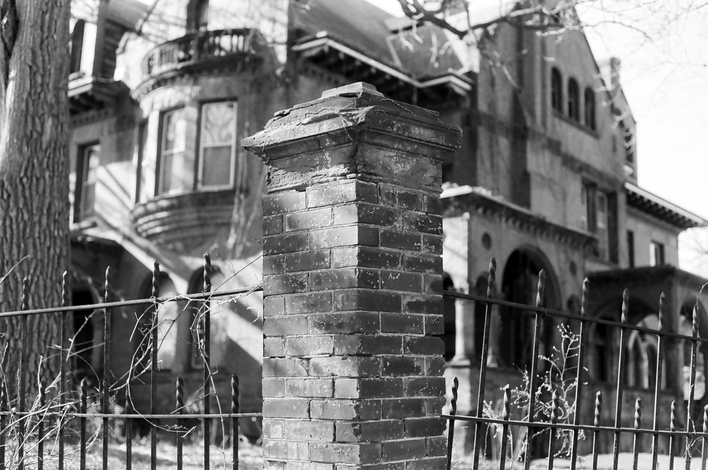 A brick pillar and wrought iron fence stand in front of a large, old house with a turret.