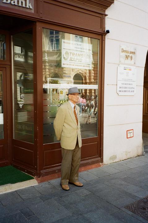 A man in a suit and hat stands on a sidewalk in front of a bank.