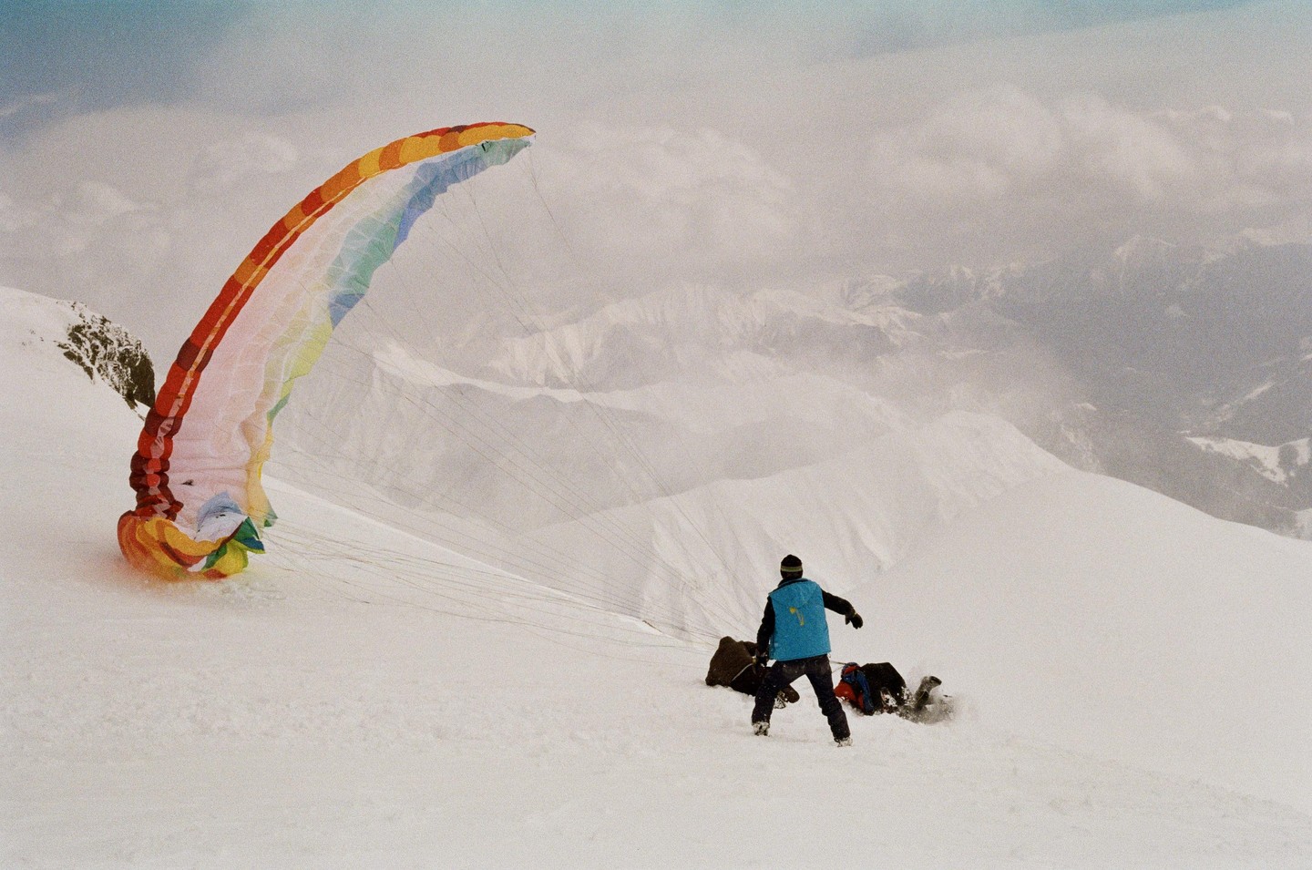 A person in a blue vest stands on a snowy mountain next to a colorful paraglider and another person on the ground.