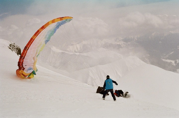 A person in a blue vest stands on a snowy mountain next to a colorful paraglider and another person on the ground.