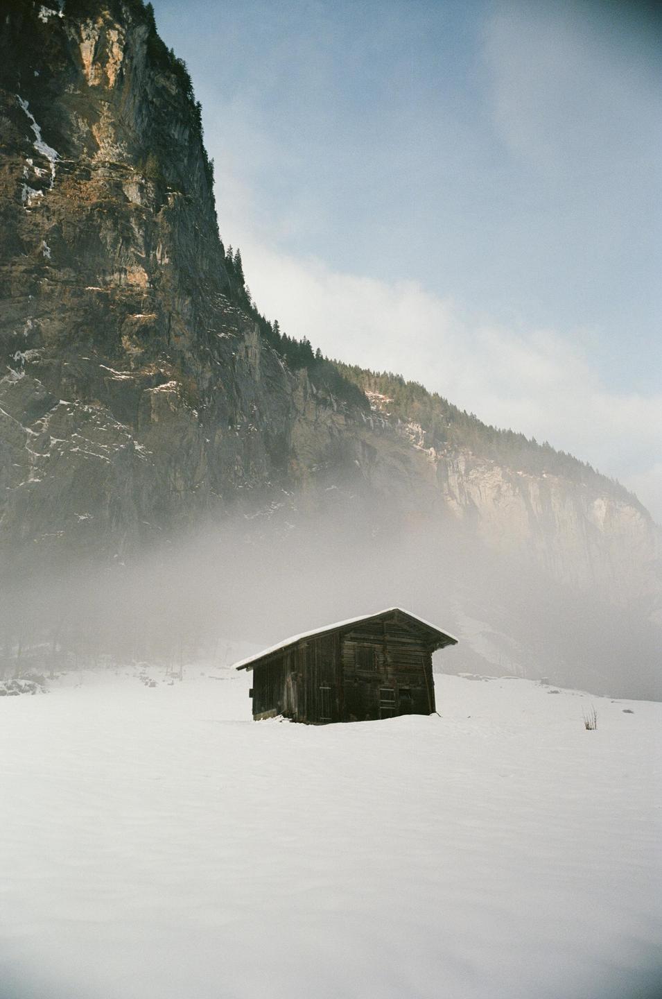 A wooden cabin sits in a snow-covered field with a large mountain and fog in the background.