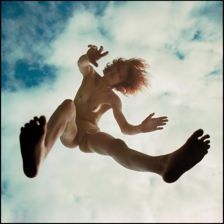 A nude woman with red hair is captured from below against a cloudy sky, appearing to fall or fly.