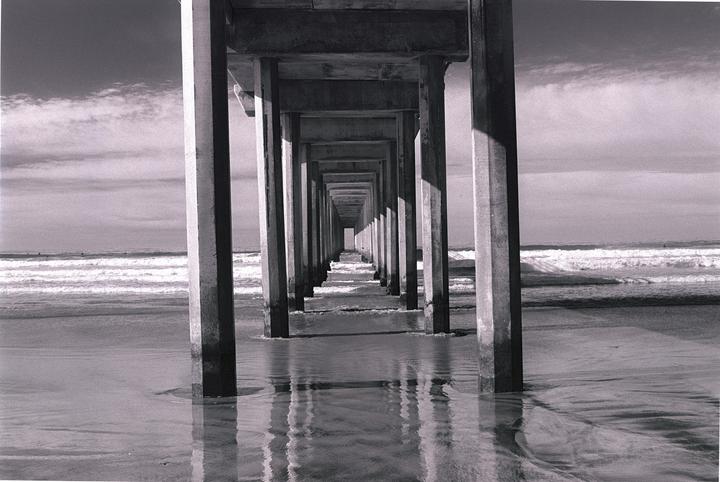 The underside of a pier with many concrete pillars extends into the distance over a sandy beach with waves.