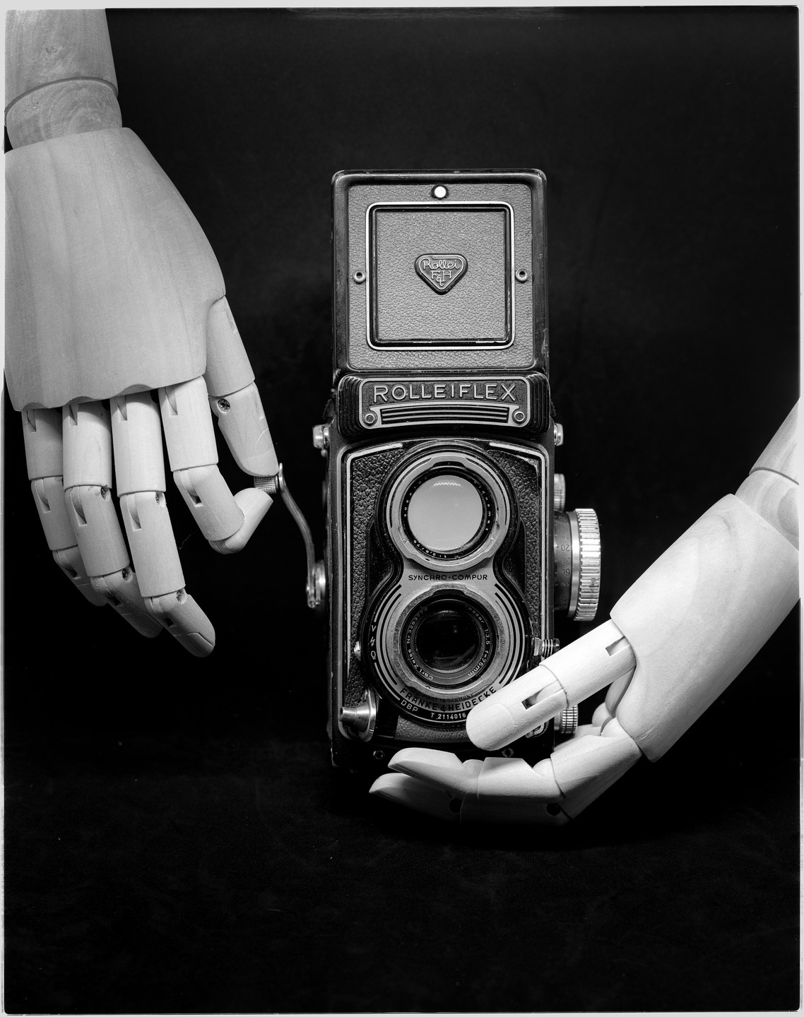 Two wooden mannequin hands hold a vintage camera against a dark background.