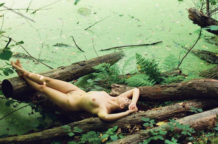 A nude woman lies on logs next to a pond covered in green algae.
