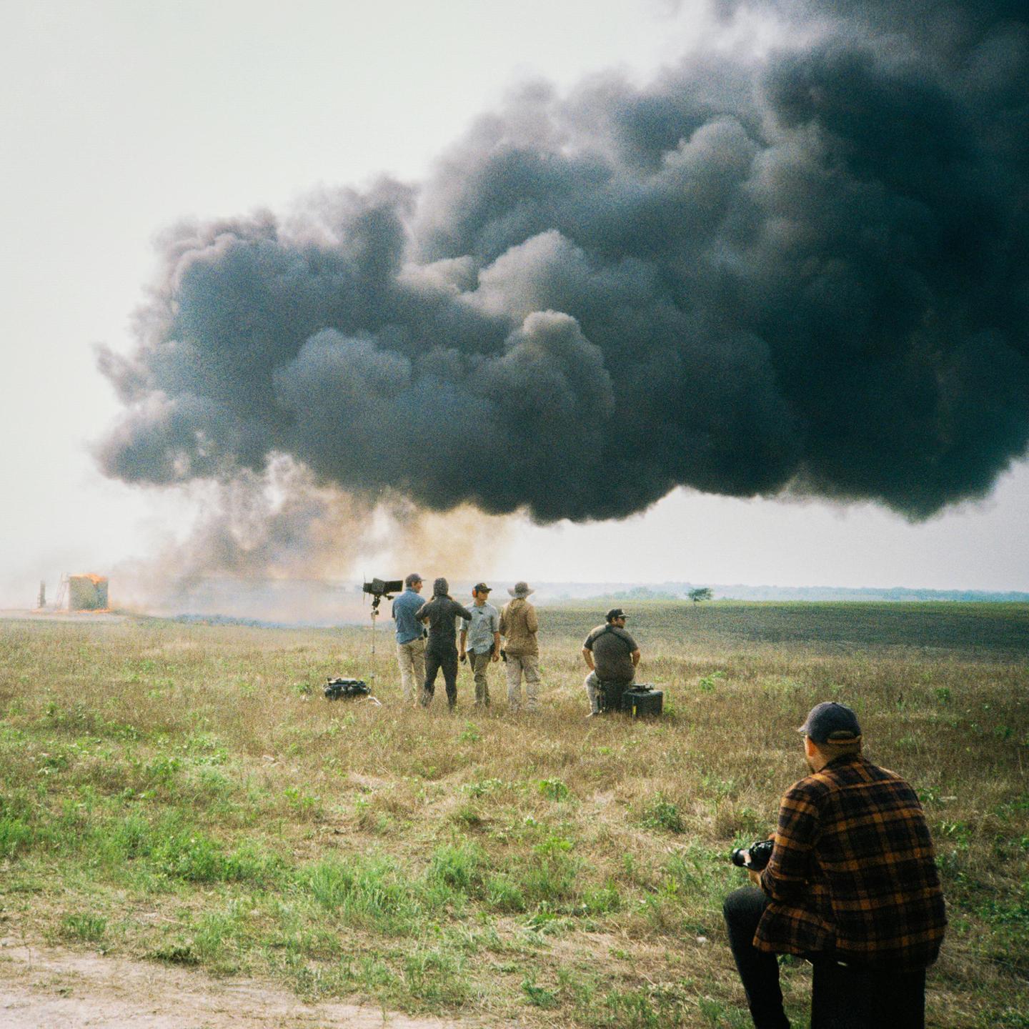A group of men are gathered in a field watching a large plume of black smoke rise from a fire in the distance.