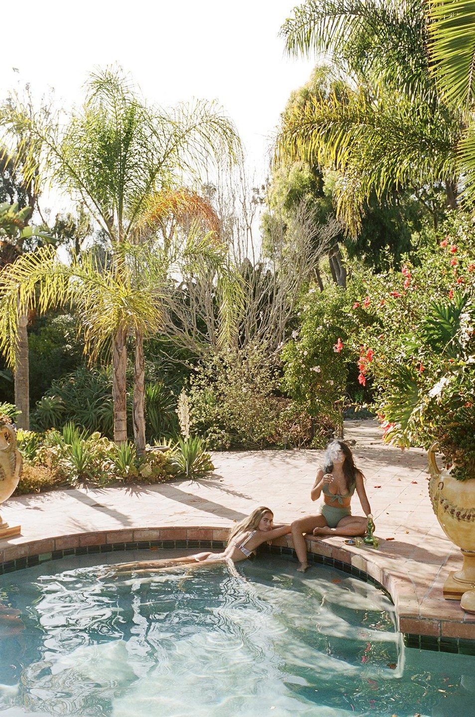 Two women in swimsuits relax by a pool surrounded by lush greenery and palm trees.