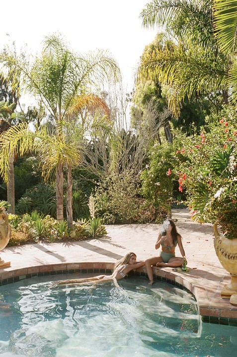Two women in swimsuits relax by a pool surrounded by lush greenery and palm trees.