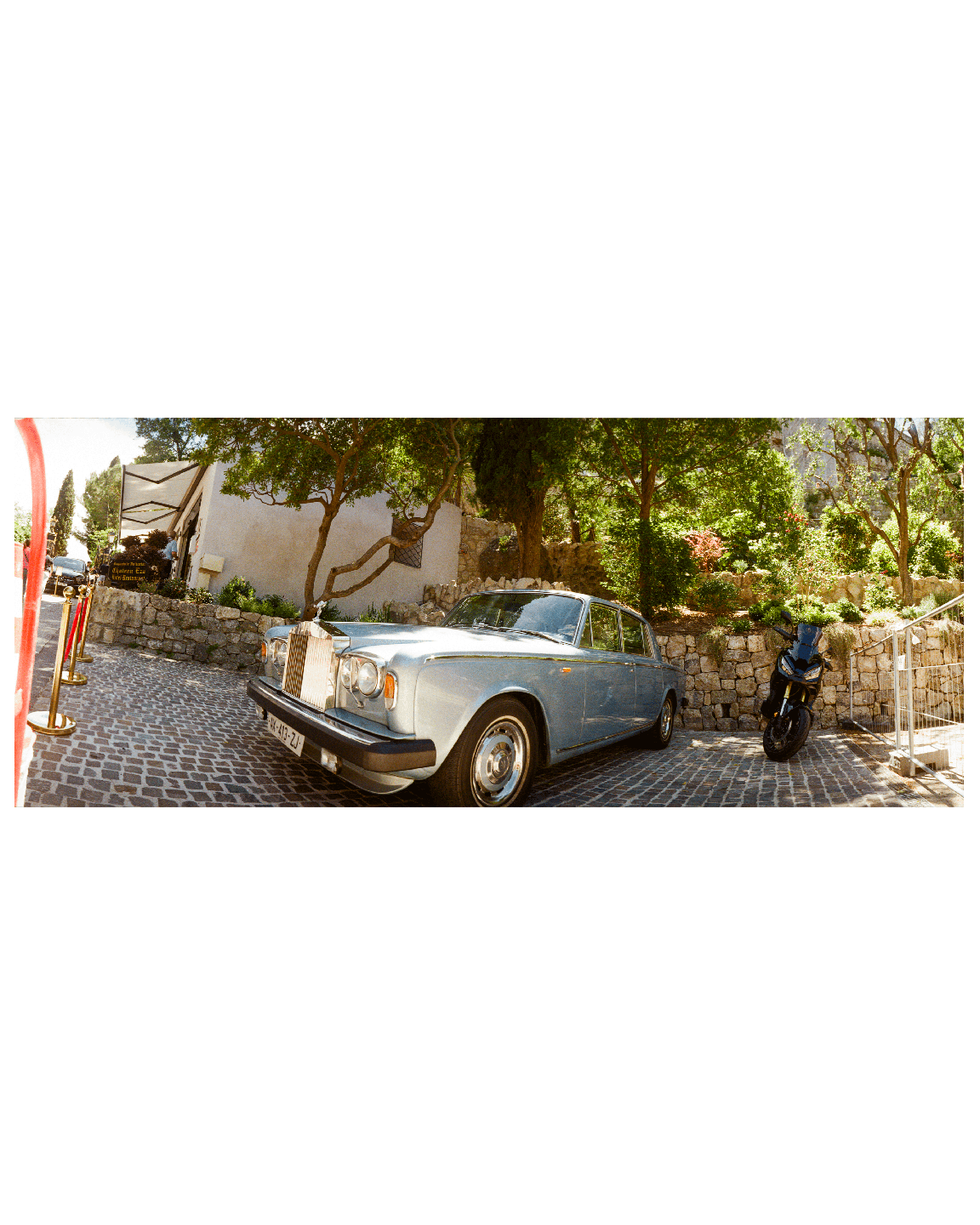 A light blue vintage car is parked on a cobblestone street next to a motorcycle and a building with trees.
