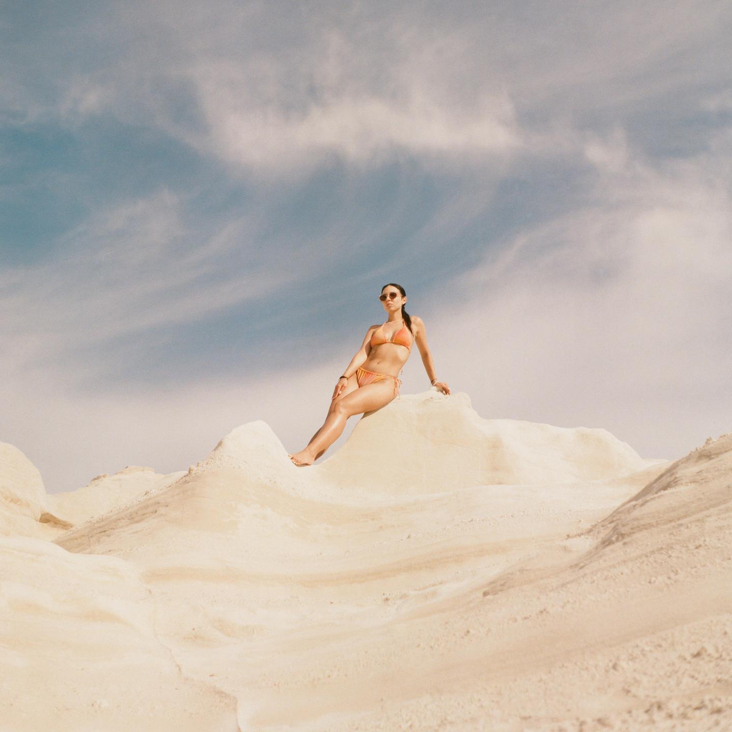 A woman in a bikini sits on a white rock formation under a blue sky with clouds.