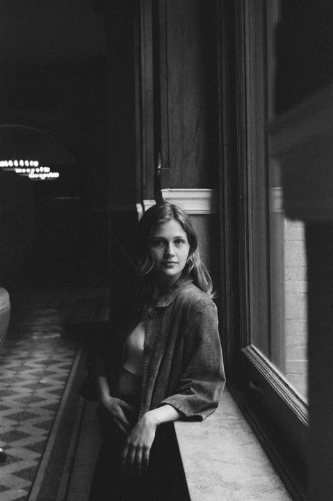 A woman with long hair leans on a window sill in a dark room.