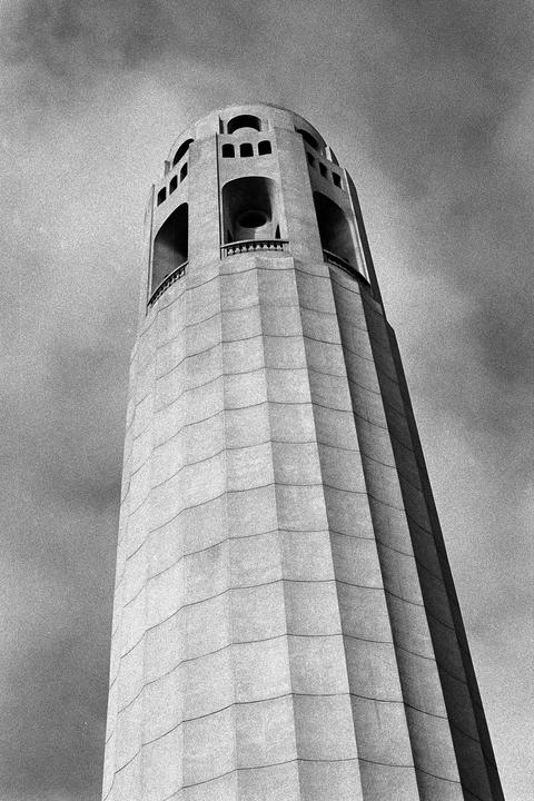 A low-angle, black and white shot captures the Coit Tower against a cloudy sky.