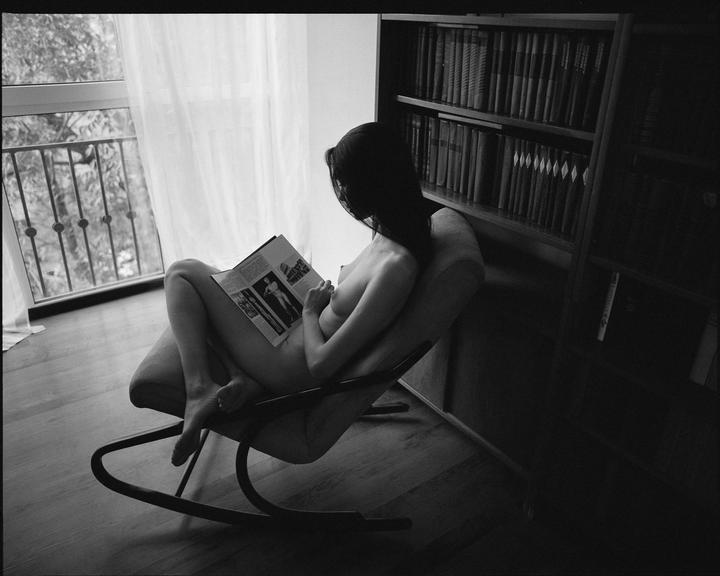A woman sits nude in a rocking chair, reading a magazine next to a window and a bookshelf.