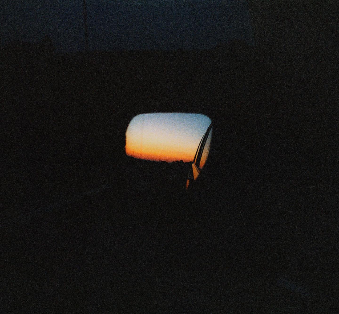 A car side mirror reflects a sunrise over a dark landscape.