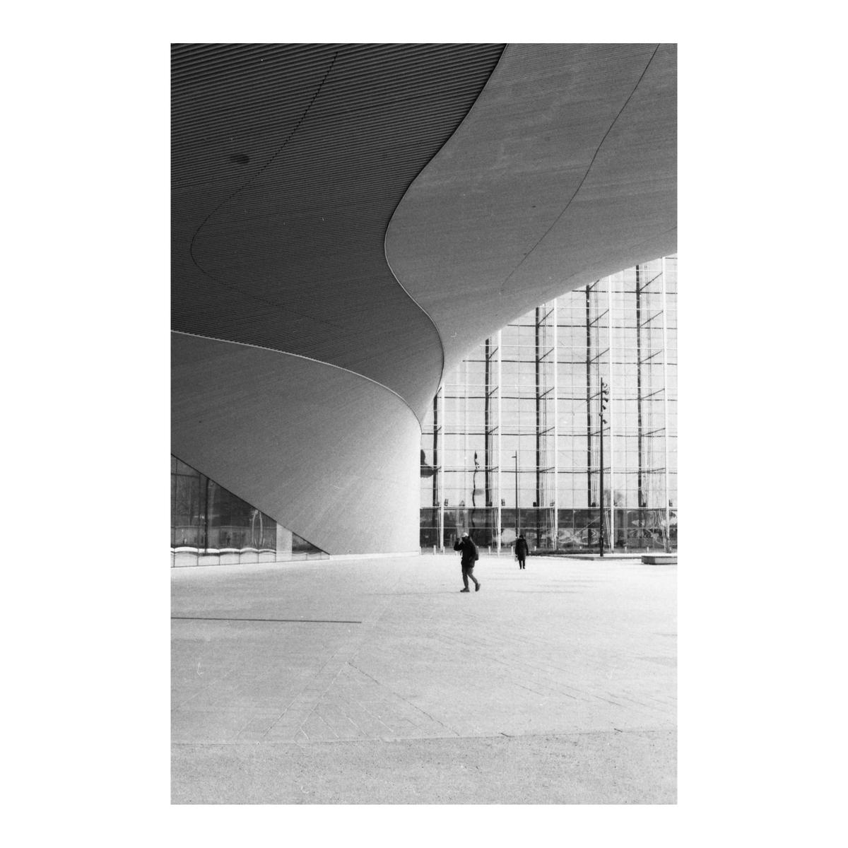 Two people walk on a paved area next to a modern building with a curved wall and a glass facade.
