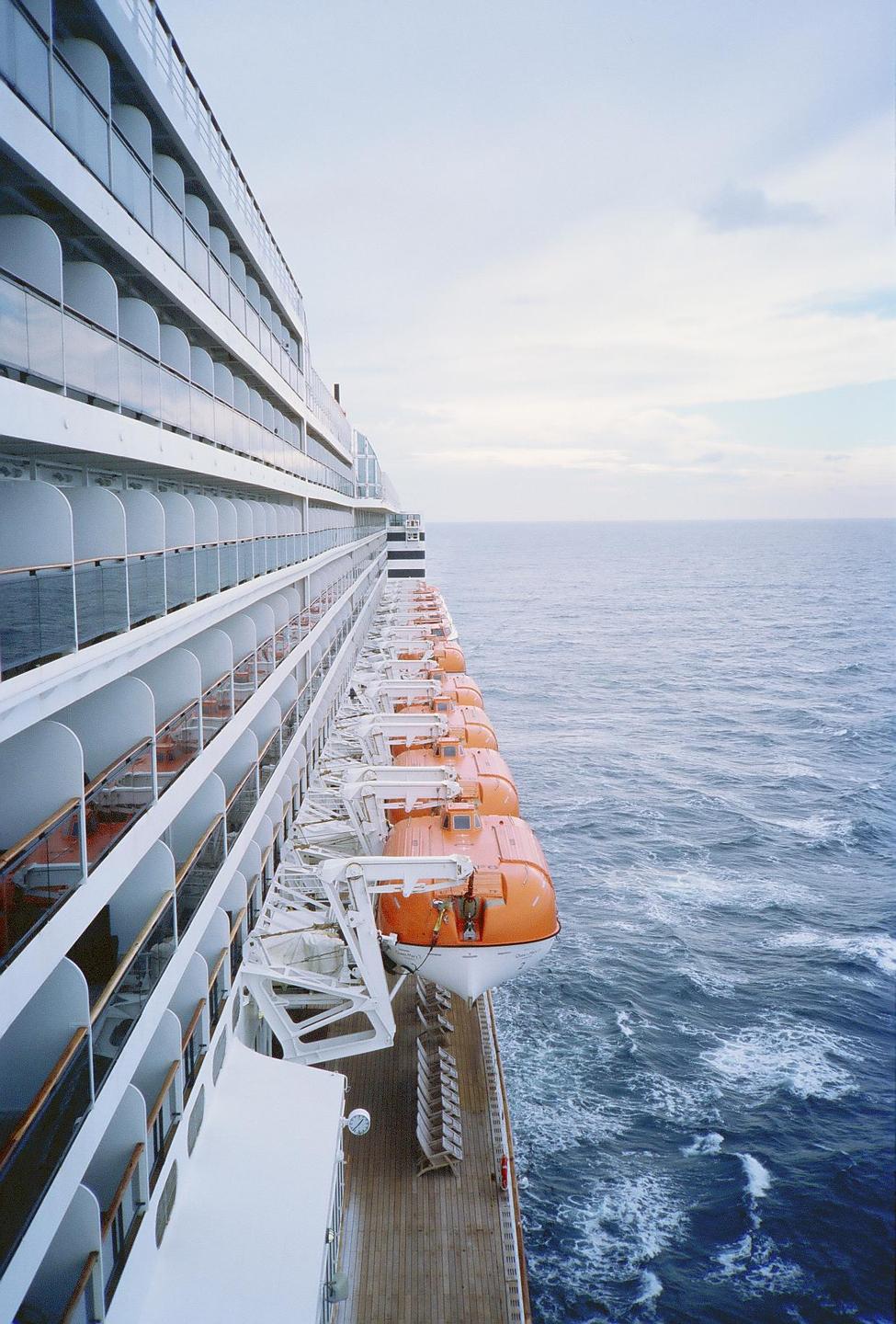 A cruise ship with many orange lifeboats is sailing on the open ocean under a cloudy sky.