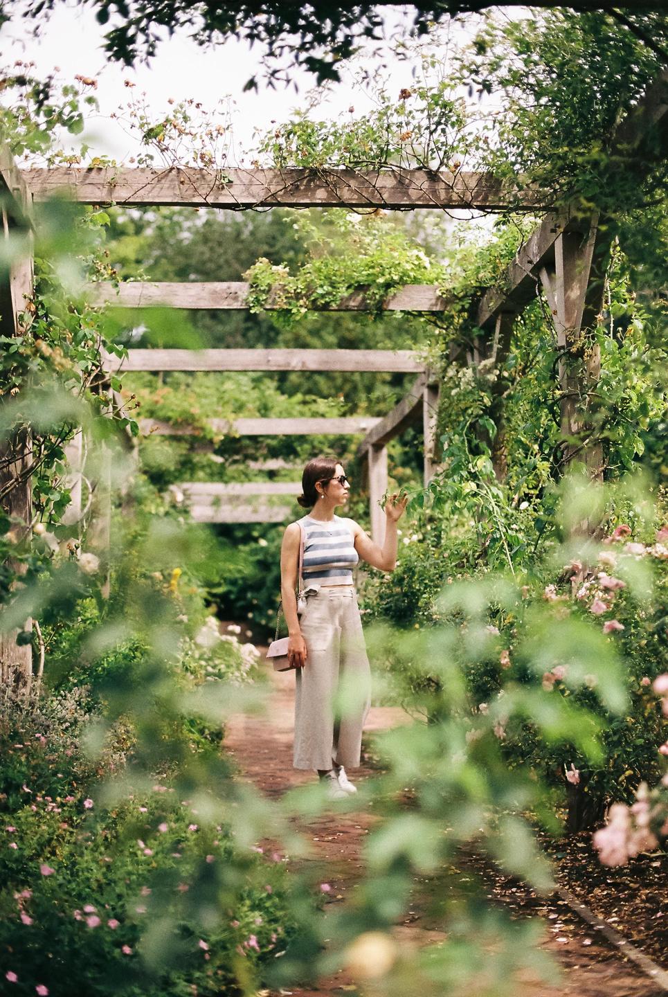 A woman stands in a garden path under a wooden pergola covered in vines and flowers.
