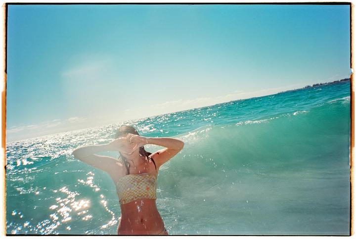 A woman in a bikini stands in the ocean with a wave breaking behind her.